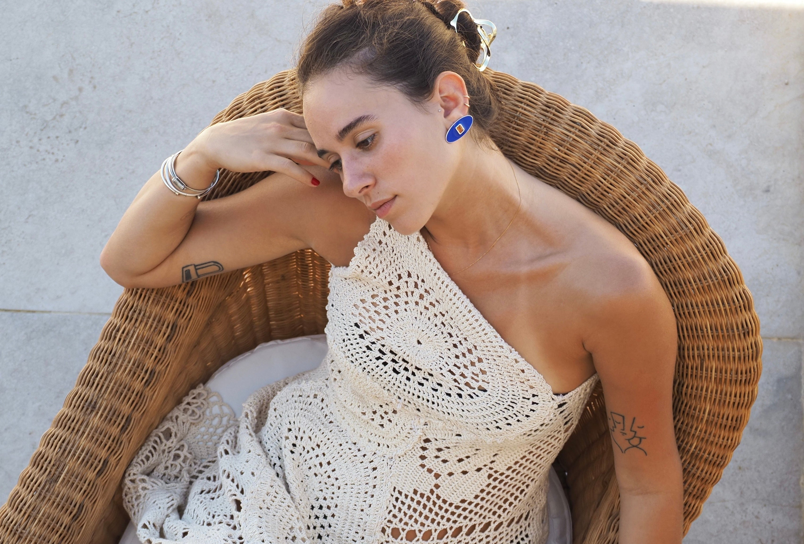 Woman sitting on a wicker chair wearing a white crochet top with a neutral background wearing enve bleu earrings